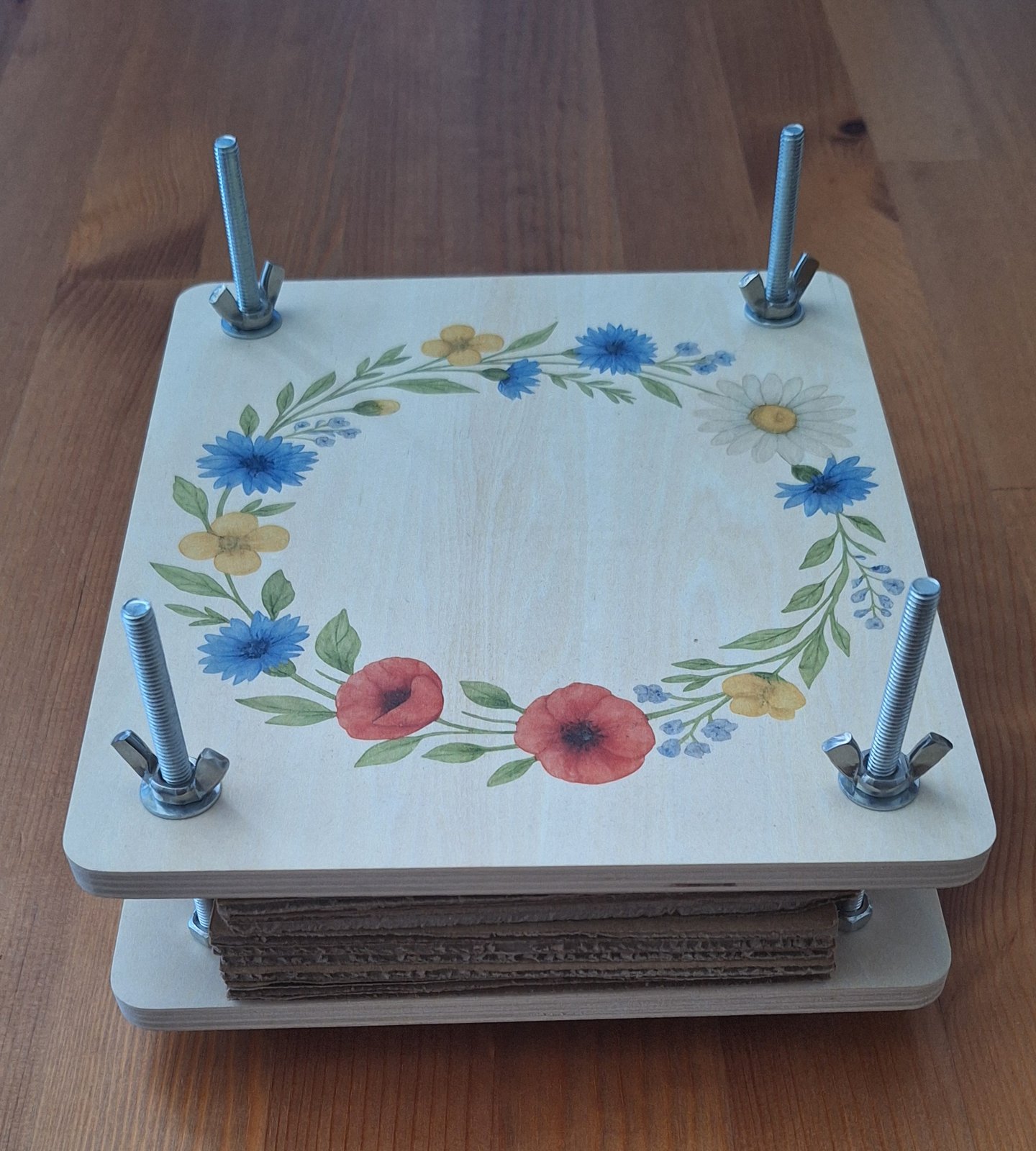 Aufgebaute Blumenpresse aus hellem Holz auf einem Holztisch. Die obere Platte trägt einen gedruckten Kranz aus Wildblumen, zwischen den Platten liegen die Kartoneinlagen.