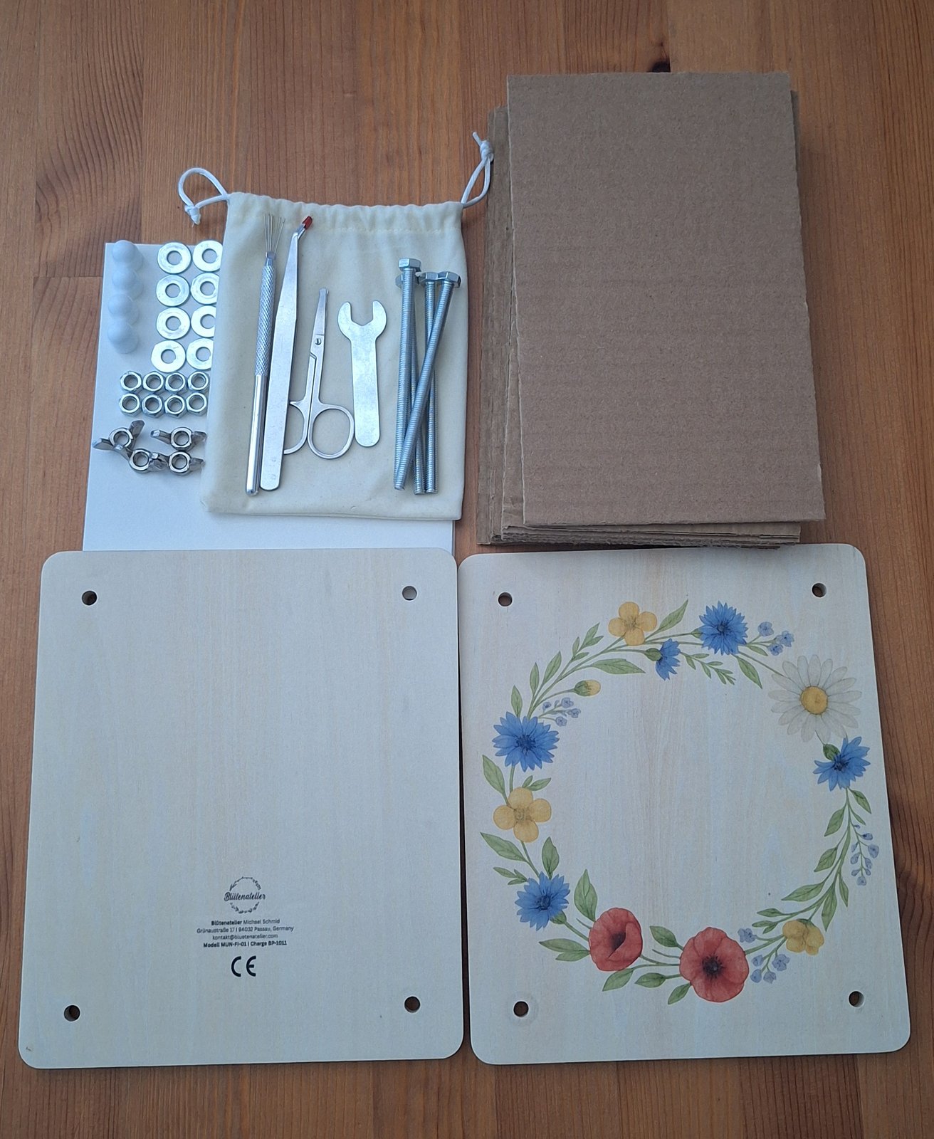 Der Lieferumfang der Blumenpresse auf einem Holztisch ausgelegt: zwei Holzplatten, ein Stapel Kartoneinlagen, Schrauben mit Flügelmuttern, Unterlegscheiben und Muttern sowie der Zubehörbeutel mit Schere, Pinzette, Nadel-Werkzeug und Maulschlüssel.